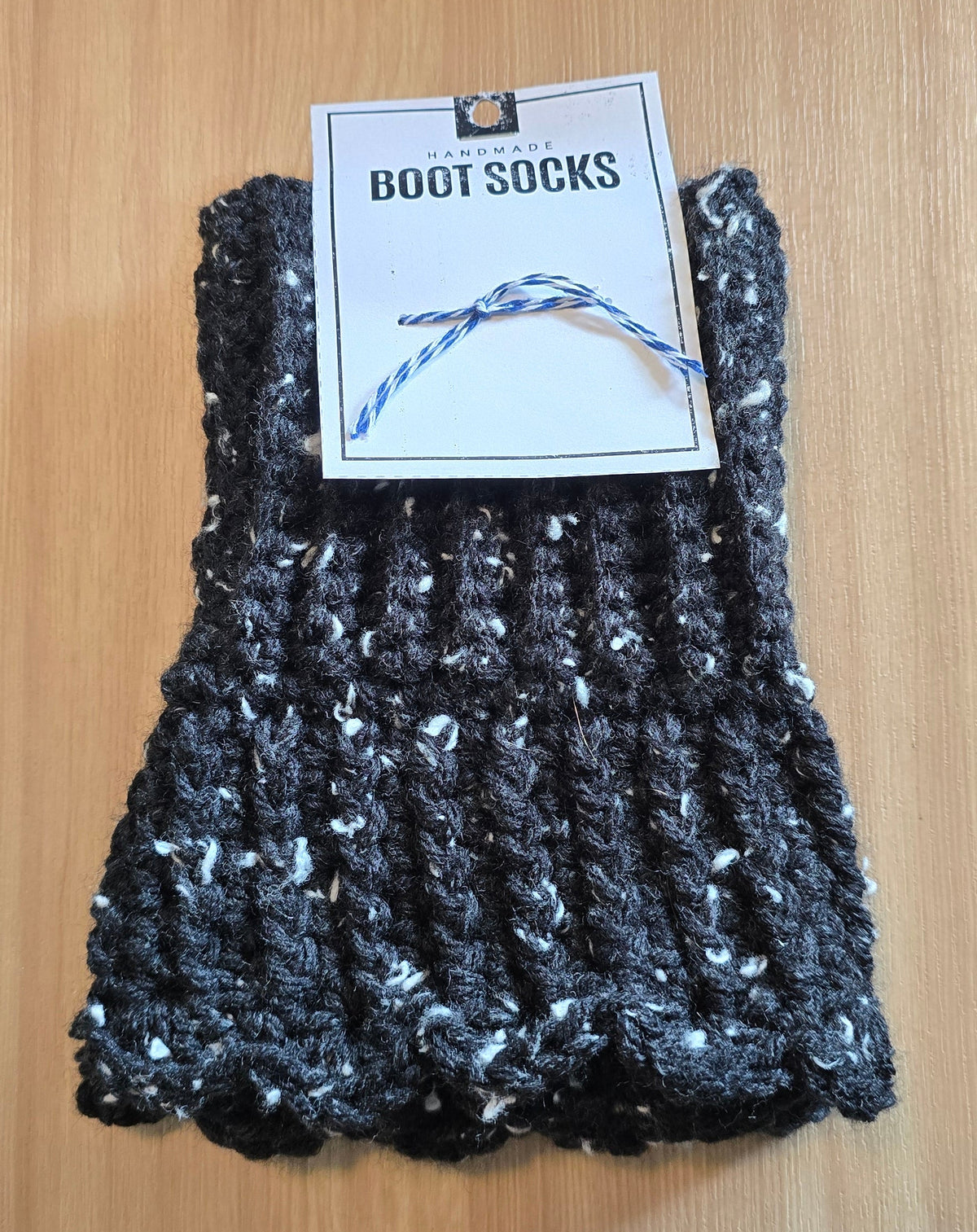 A pair of handmade black and white marled chunky knit boot socks with a white paper tag that reads 'BOOT SOCKS' on a light wood background.