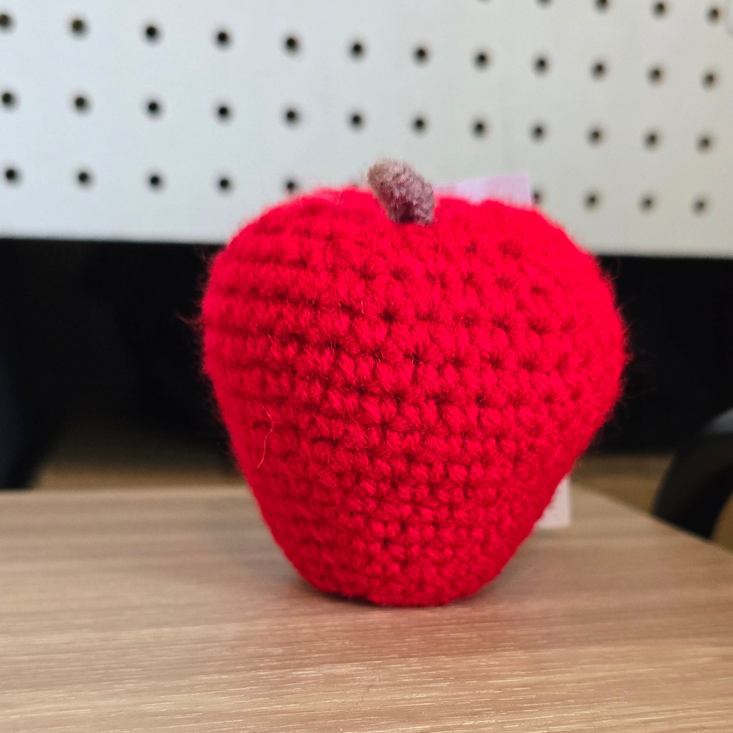 A bright red crocheted apple with a small brown stem, resting on a light wood desk in front of a white pegboard wall.