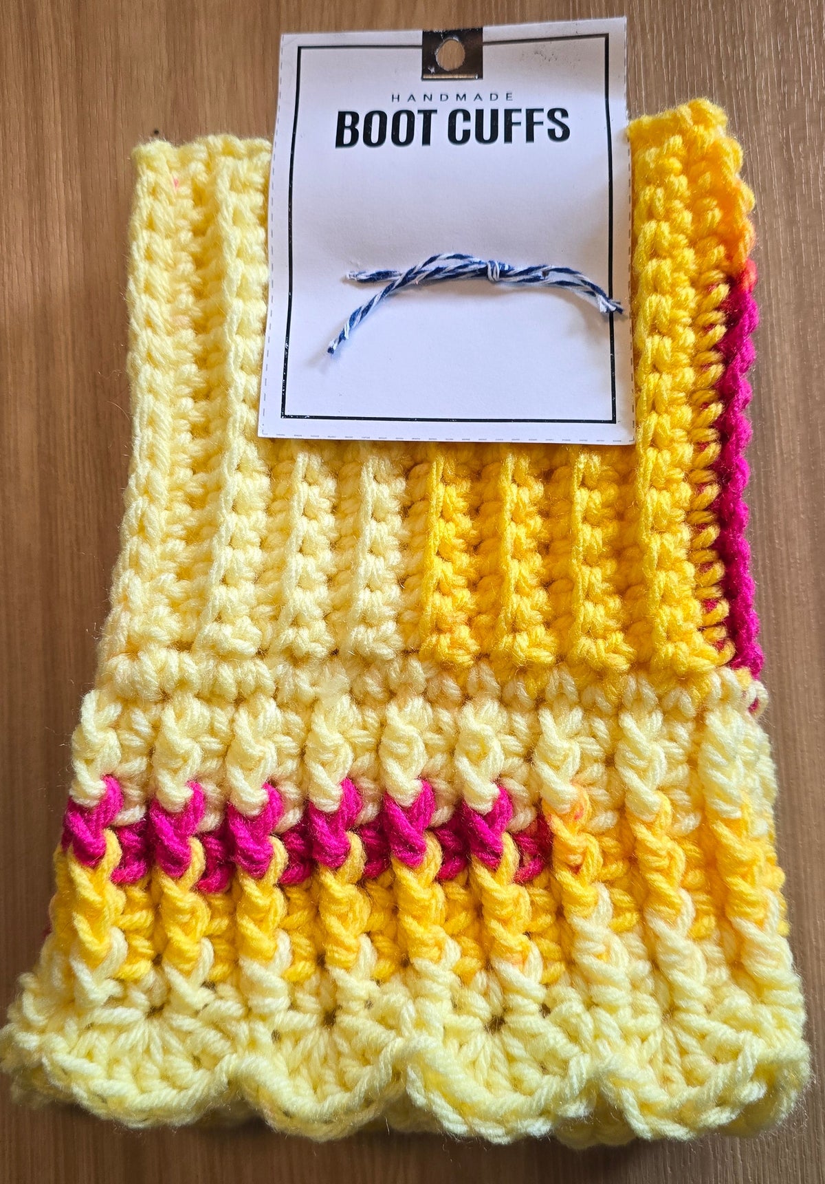 A pair of hand-crocheted boot cuffs in yellow and pink yarn, with a white paper tag attached that reads 'BOOT CUFFS'. The cuffs are laid flat on a wooden table.