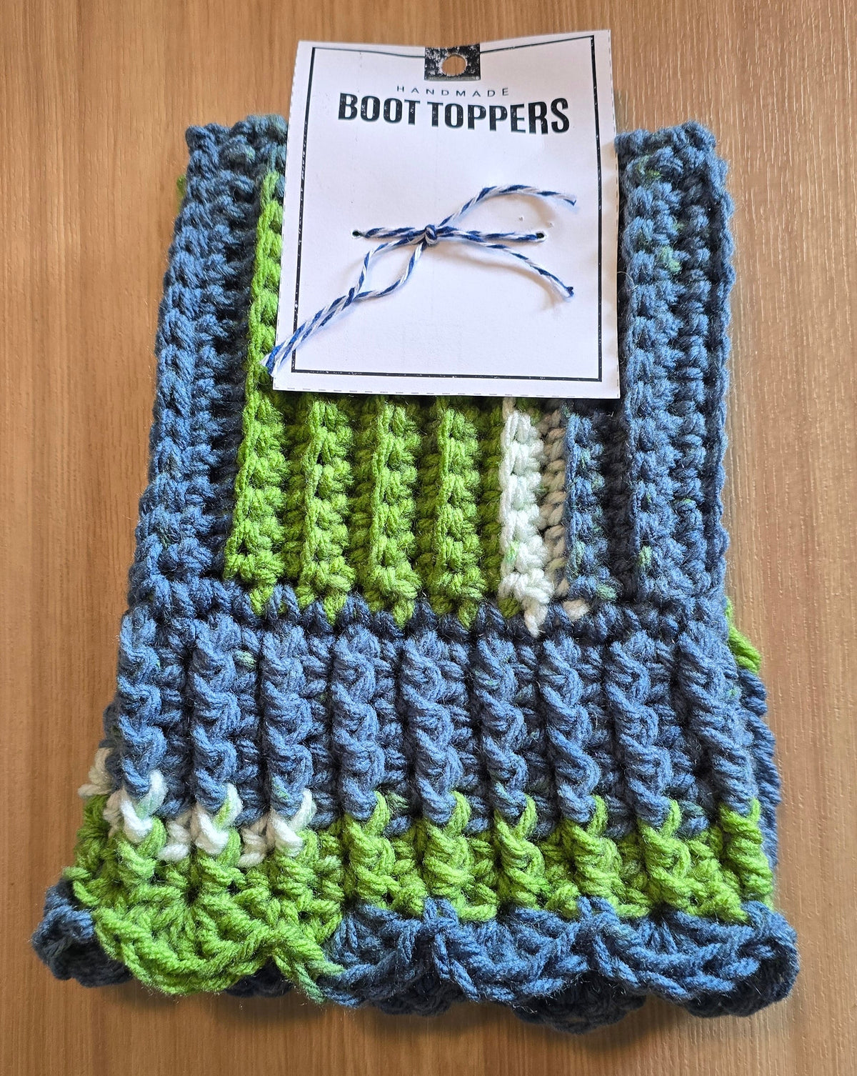 A handmade crocheted boot topper in blue, green, and white stripes, displayed on a wooden table with its paper tag attached.