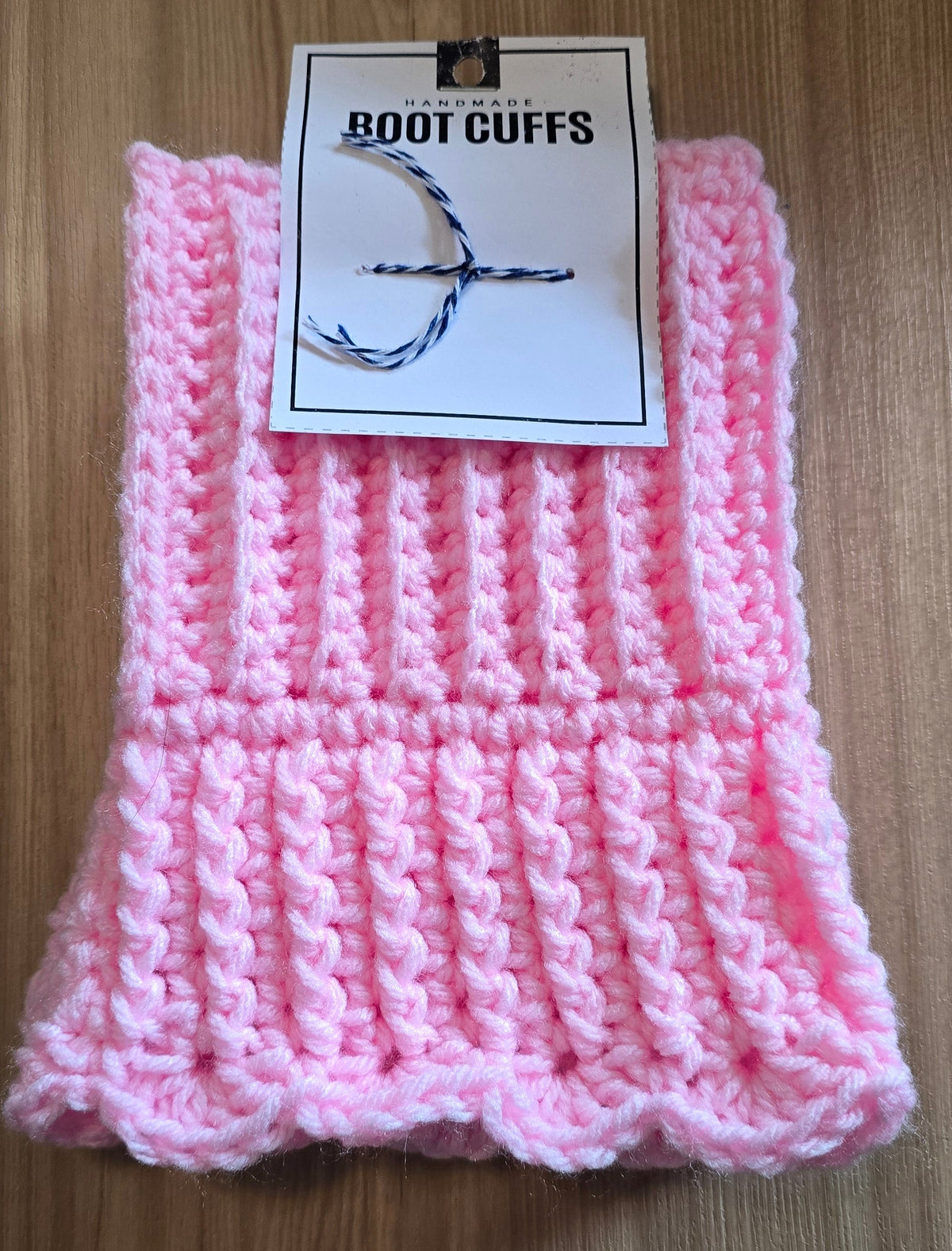A pair of pink crocheted boot cuffs with a ribbed texture and ruffled edges, shown with a paper tag that reads 'CROCHETED BOOT CUFFS' on a wooden surface.